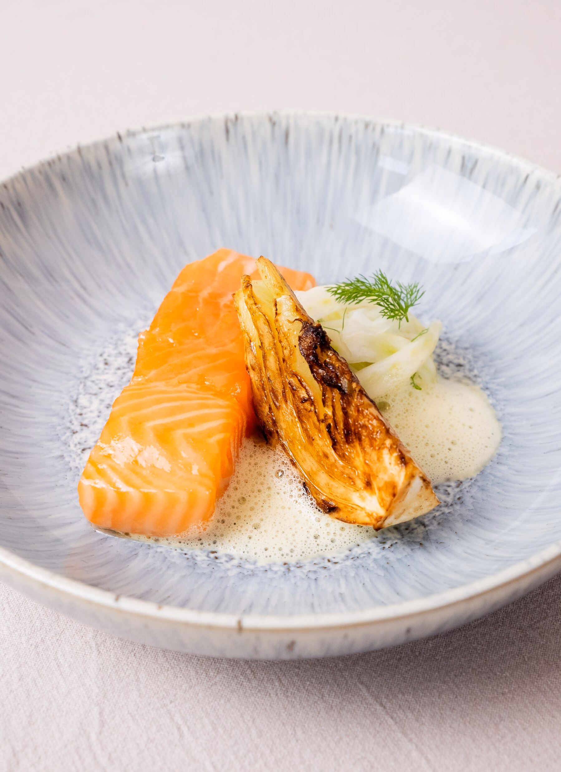Een filet zalm met een gebakken partje venkel, ingelegde venkel en een schuimige beurre blanc in een blauwgrijs aardewerken bord.