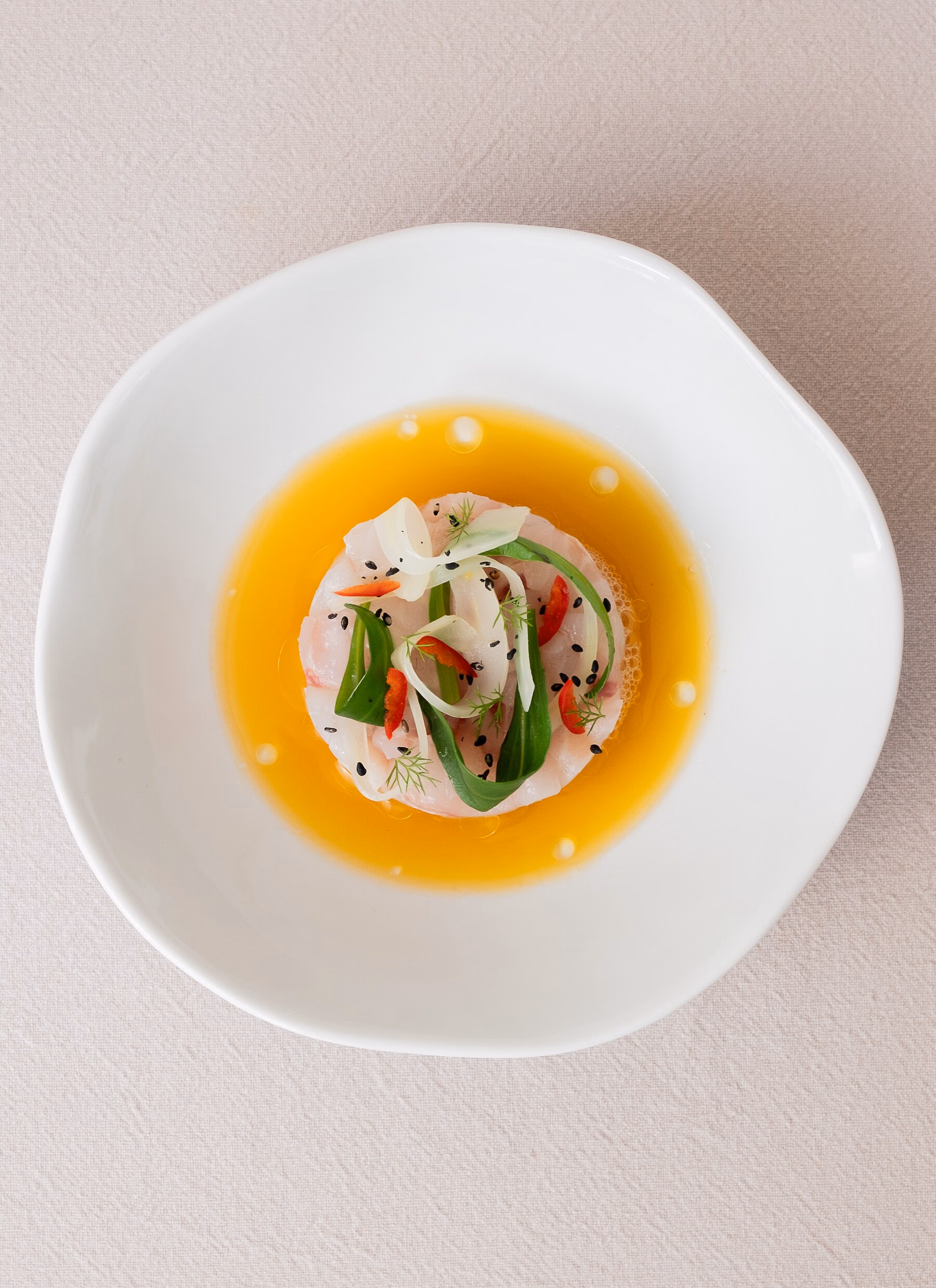 Tartaar van zeebaars met venkel, rode peper en sesamzaad, omringd door een oranje sinaasappeldressing waarin druppels sjalotolie drijven.